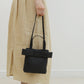 Person holding a black handbag with a bow detail, wearing a beige skirt and black shoes on a plain background.