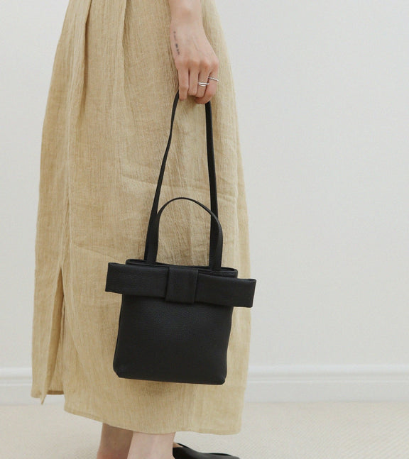 Person holding a black handbag with a bow detail, wearing a beige skirt and black shoes on a plain background.