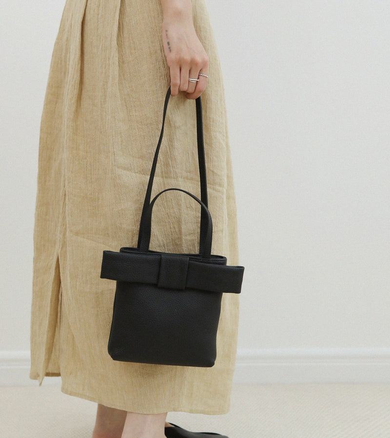 Person holding a black handbag with a bow detail, wearing a beige skirt and black shoes on a plain background.