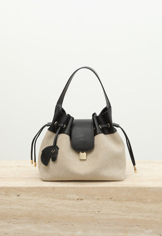 A beige canvas bucket bag with black leather trim, dual top handles, and drawstring details, displayed on a light stone surface against a white background with 'Shevennie Studio' text at the top.