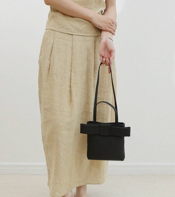 Person holding a black handbag with a bow detail against a white background, wearing a beige outfit.